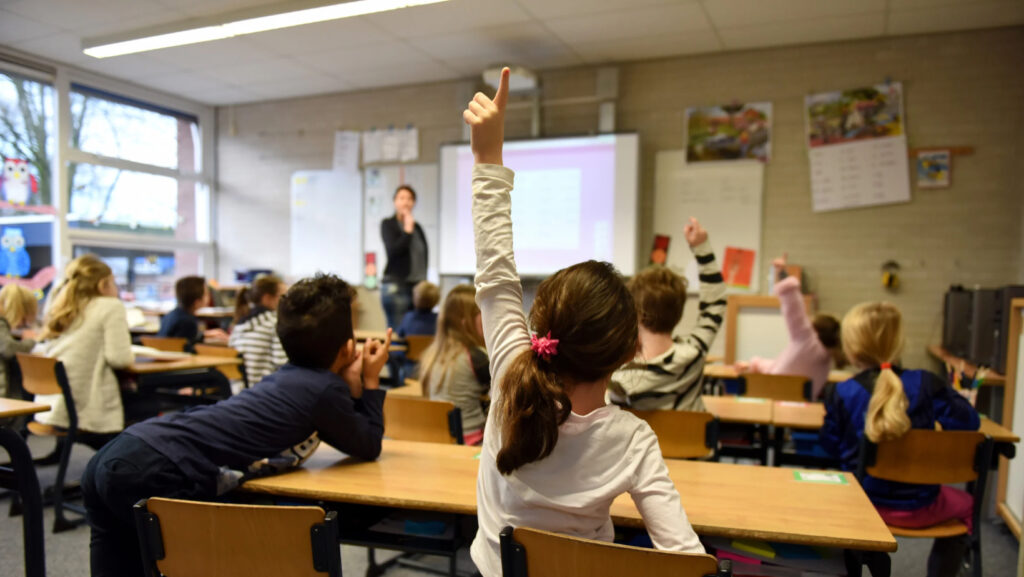 Students in a classroom, facing teacher with hands raised